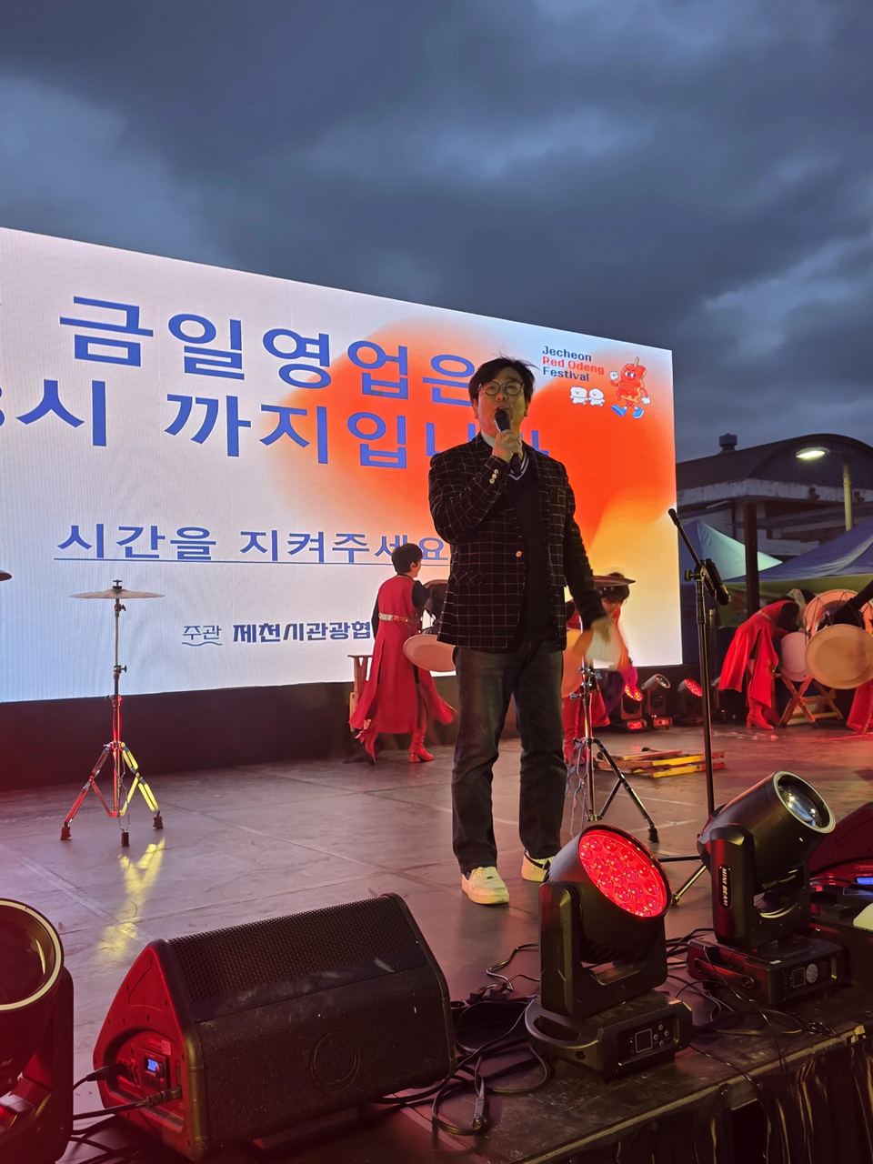 야외 무대에서 마이크를 들고 서 있는 장면 · 야외 축제 행사