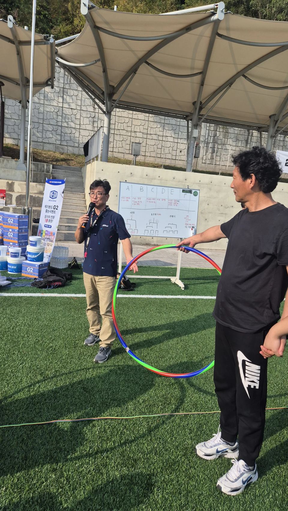 훌라후프를 든 참가자 옆에서 마이크로 진행하는 장면 · 체육 행사